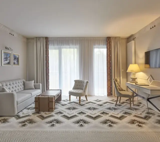Elegancki pokój w hotelu 5 gwiazdkowym: sofa, stolik, fotele, biurko i TV. Wzory na dywanie i zasłonach dodają charakteru.