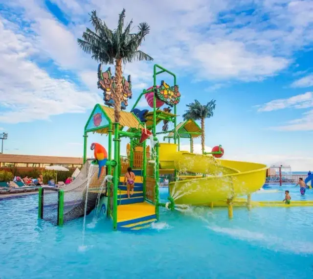 Kolorowy aquapark przy hotelu z palmami i zjeżdżalniami. Dzieci bawią się w wodzie, a na leżakach odpoczywają goście.