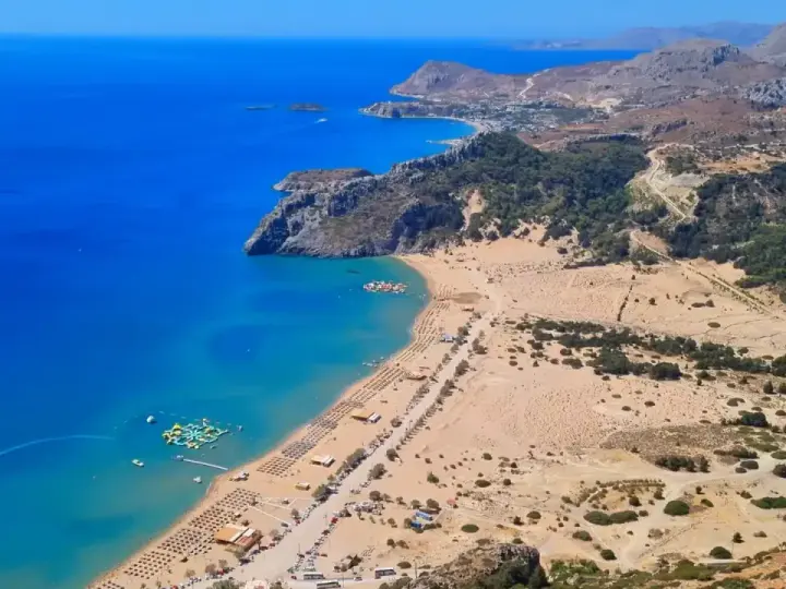 Malownicze **Rodos plaże** z turkusową wodą, piaszczystym brzegiem i zielonymi wzgórzami w tle. Widać wodny plac zabaw.