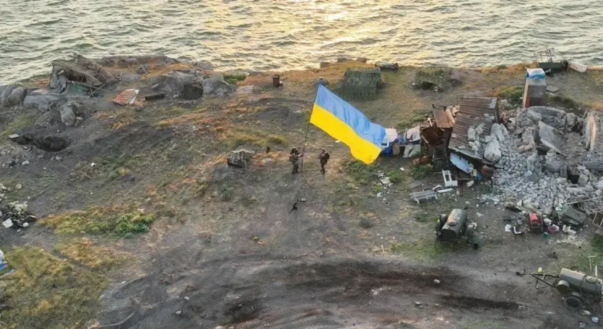 Na wyspie węży, pośród zniszczeń, żołnierze dumnie wznoszą ukraińską flagę. W tle morze i ruiny.
