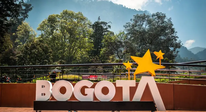 Napis BOGOTA z żółtymi gwiazdami, symbolizujący stolicę Kolumbii, na tle zielonych wzgórz i błękitnego nieba.