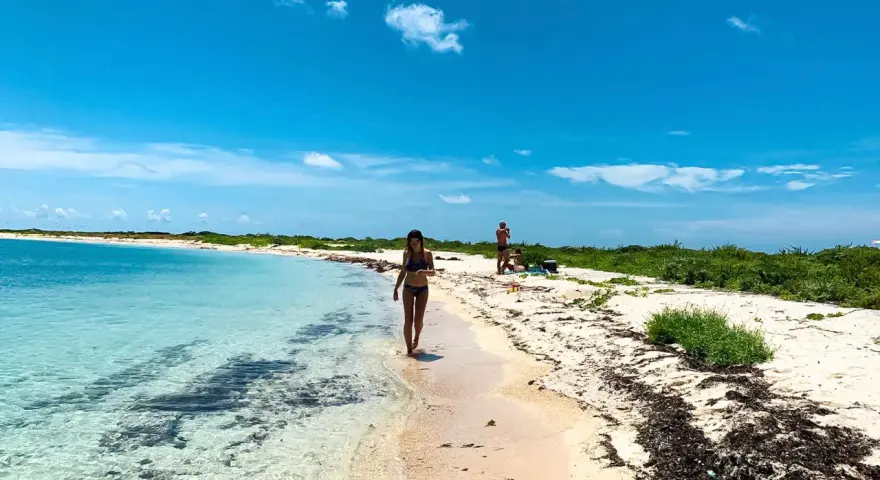 Kobieta spaceruje po piaszczystej plaży nad turkusową wodą. W tle widać innych ludzi i bujną zieleń. To idealne miejsce na wypoczynek nad Zatoką Meksykańską.
