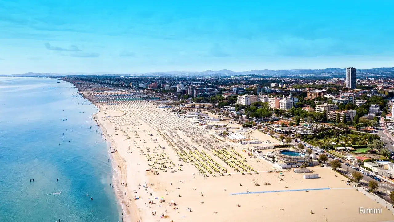 Słoneczna plaża w Rimini, pełna leżaków i parasoli, z turkusowym morzem i miastem w tle.