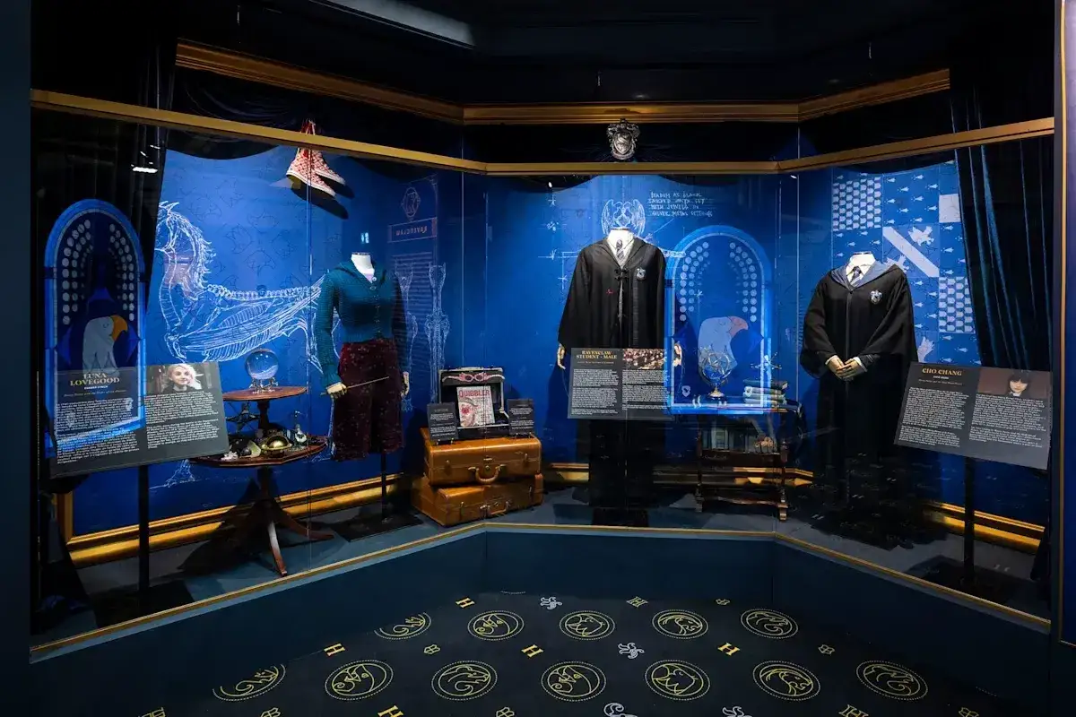 Wystawa w muzeum Harry'ego Pottera prezentuje stroje i rekwizyty postaci, w tym Luny Lovegood i Cho Chang, na tle szkiców i herbów Hogwartu.