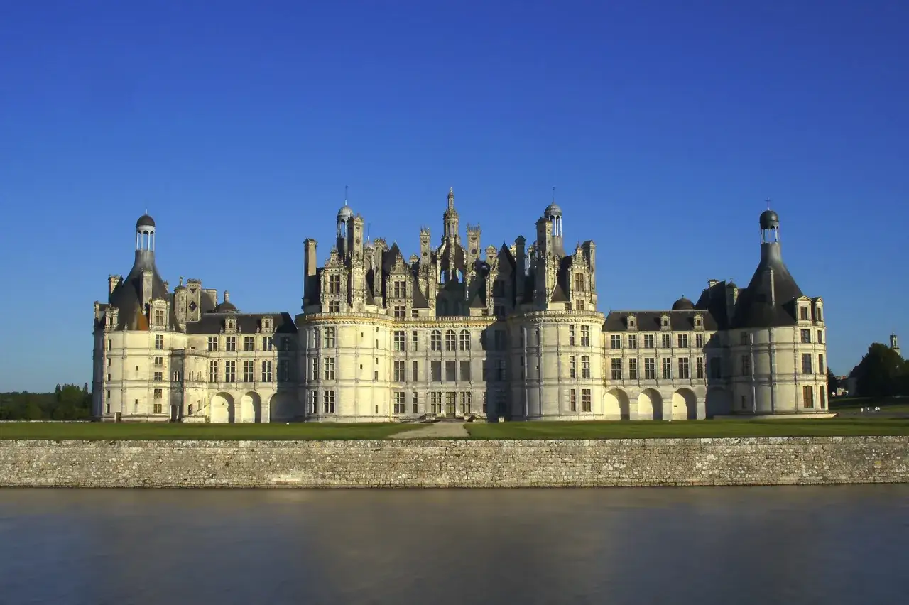 Zamek Chambord nad spokojną wodą, przypominającą najdłuższą rzekę Francji. Majestatyczna budowla z licznymi wieżyczkami i oknami.