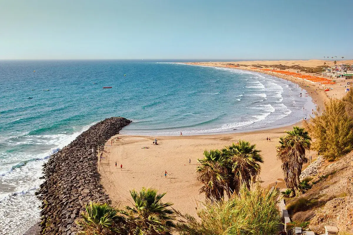 Słoneczna plaża Playa del Inglés z turkusową wodą, kamiennym falochronem i palmami.