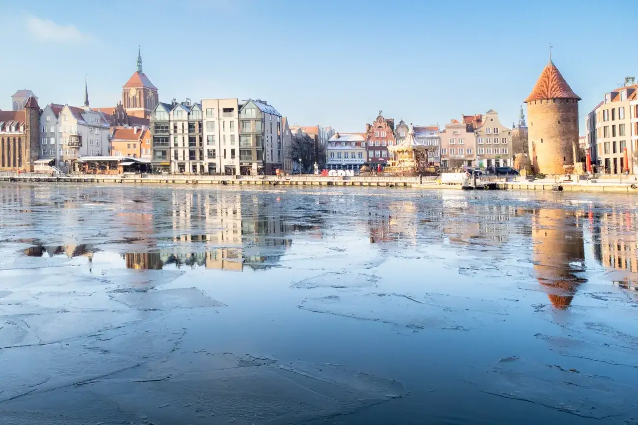 Zamarznięta rzeka na m. Gdańsk, z widokiem na zabytkową zabudowę i wieżę.