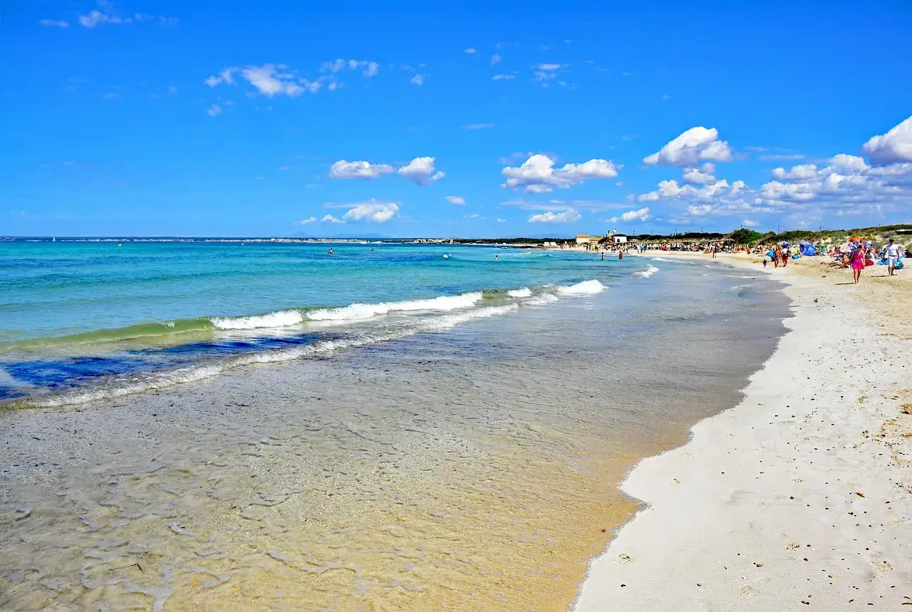 Piękna plaża Es Trenc z turkusową wodą i białym piaskiem, pełna wypoczywających ludzi pod błękitnym niebem z chmurkami.