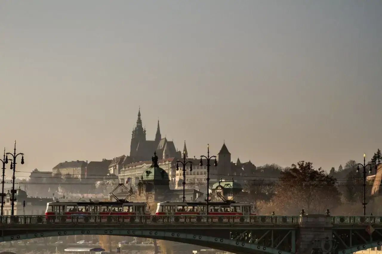 Tram jedzie przez most w Czechach, w tle widać zamek i zabytkowe budynki.