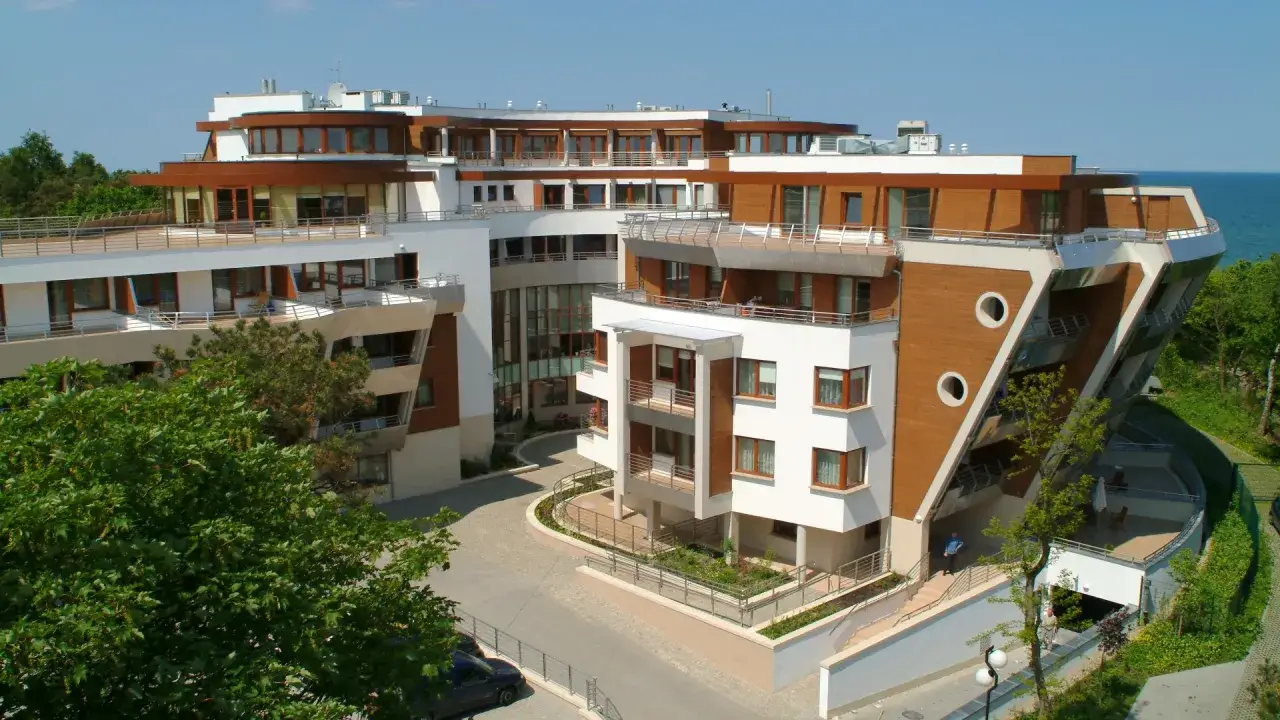 Nowoczesny hotel 5 gwiazdkowy nad morzem, z białymi fasadami i drewnianymi elementami, otoczony zielenią.