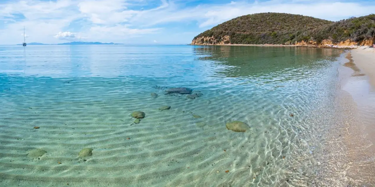 Piękne livorno plaże z krystalicznie czystą wodą i piaszczystym dnem. W oddali widać żaglówkę i zielone wzgórze.