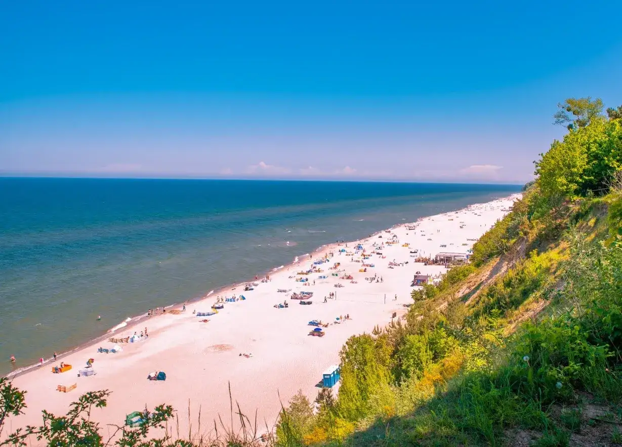 Idealne miejsce, gdzie na wakacje w lipcu: szeroka, piaszczysta plaża nad spokojnym morzem, pełna wypoczywających ludzi i zielone klify.