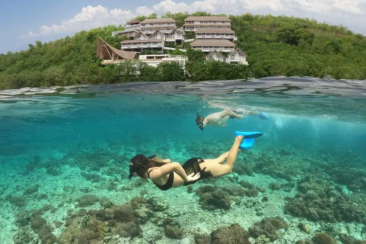 Para nurkuje w krystalicznie czystej wodzie, podziwiając rafy koralowe. Nad nimi, na zielonym wzgórzu, widać luksusowy hotel, idealne miejsce na wakacje na Bali.