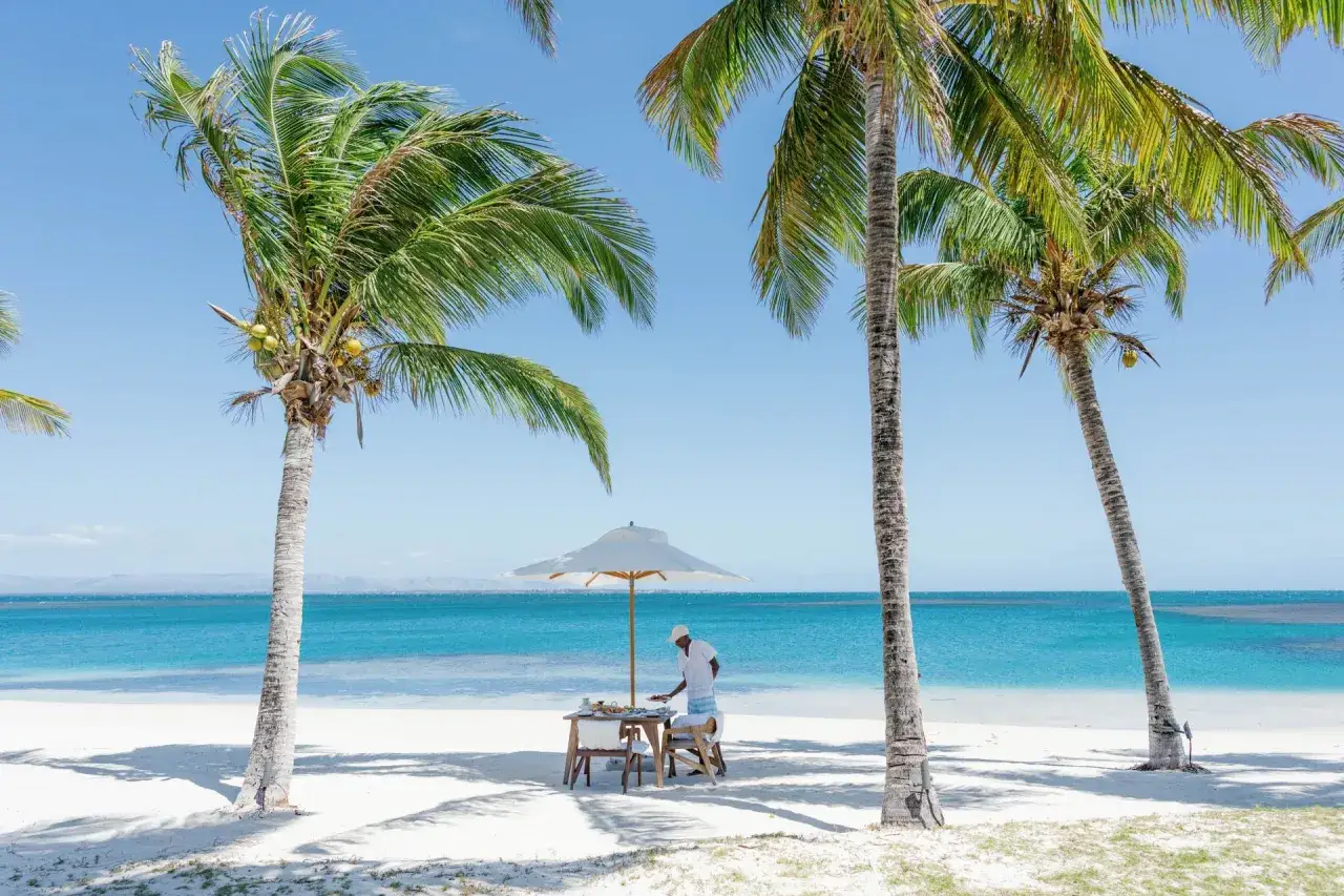 Idealne wakacje na Madagaskarze: obsługa przygotowuje stolik pod palmami na plaży, z widokiem na turkusowe morze.