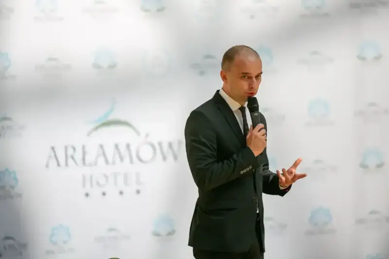 Mężczyzna w garniturze przemawia na tle logo Hotel Arłamów. Właściciel hotelu opowiada o swoich sukcesach.
