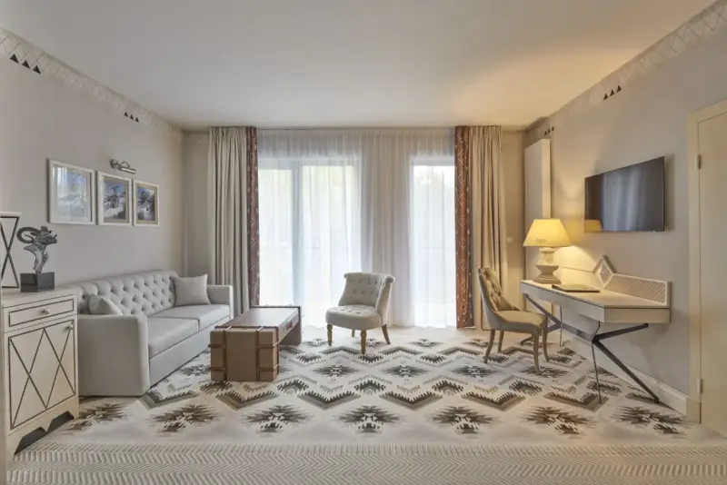 Elegancki pokój w hotelu 5 gwiazdkowym: sofa, stolik, fotele, biurko i TV. Wzory na dywanie i zasłonach dodają charakteru.
