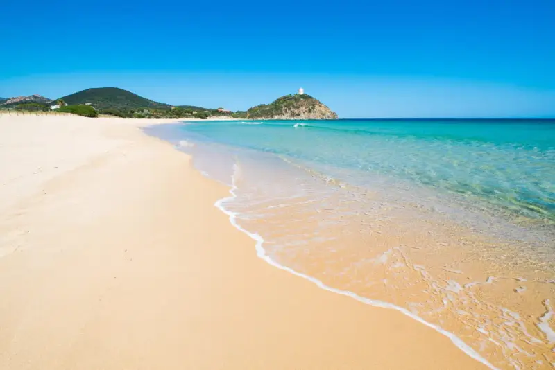 Piękne plaże Cagliari z turkusową wodą i piaszczystym brzegiem. W oddali widać wzgórze z wieżą.