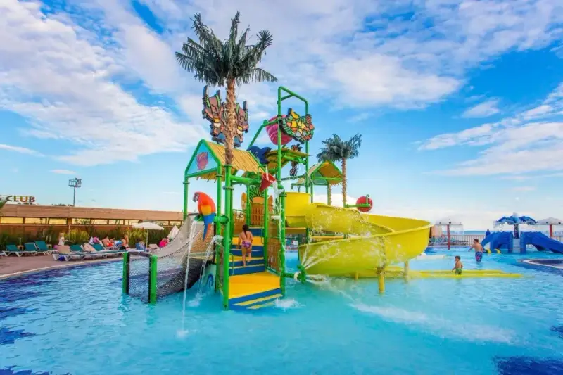 Kolorowy aquapark przy hotelu z palmami i zjeżdżalniami. Dzieci bawią się w wodzie, a na leżakach odpoczywają goście.